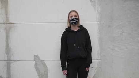 Young-Female-in-Black-Clothes-and-Face-Mask-Standing-by-Wall-Looking-at-Camera