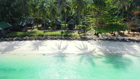 Beach-cabins-under-palm-trees-with-green-leaves-shade-over-white-sand-of-exotic-beach-washed-by-calm-turquoise-lagoon