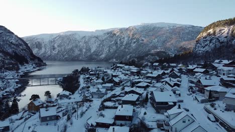 Flying-close-to-houses-in-snowy-town-Stanghelle---Forward-moving-aerial-against-fjord,-mountain,-bridge-and-sun