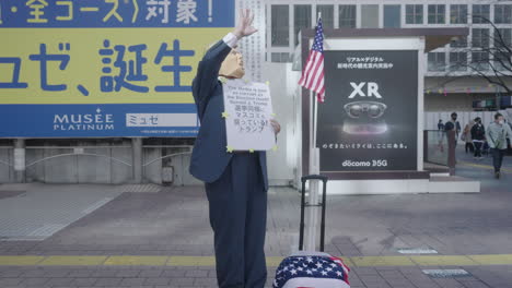 Hombre-Con-Máscara-Y-Vestido-Como-Donald-Trump-De-Pie-Con-Pancarta-Y-Levantando-La-Mano-En-La-Plaza-Hachiko-Durante-La-Pandemia-En-Tokio,-Japón