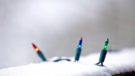 Nahaufnahme-Von-Blauen,-Grünen-Und-Orangefarbenen-Weihnachtslichtern-Auf-Einem-Verschneiten-Deck-Mit-Verschwommenem-Hintergrund-Und-Fallendem-Schnee