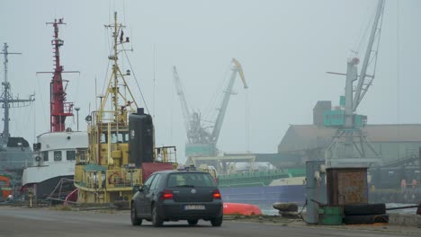Port-cranes-loading-dry-cargo-ship-at-Port-of-Liepaja-in-foggy-day,-car-passes-by,-wide-shot