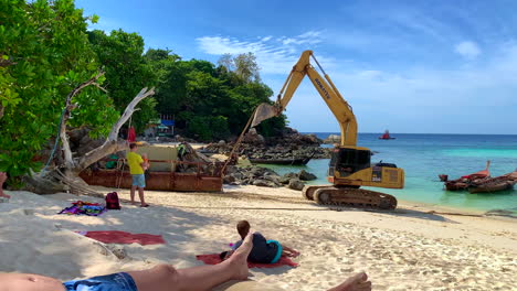 A-shot-of-an-excavator-dragging-a-petrol-or-oil-tank-by-the-beach-with-men-working-around-it-and-tourists-watching-on-the-side