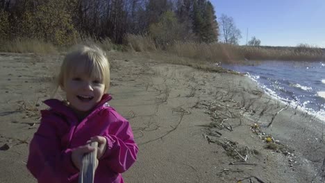 Cute-and-happy-Caucasian-girl,-three-years-old,-talking-and-playing-with-stick-at-beach-in-autumn