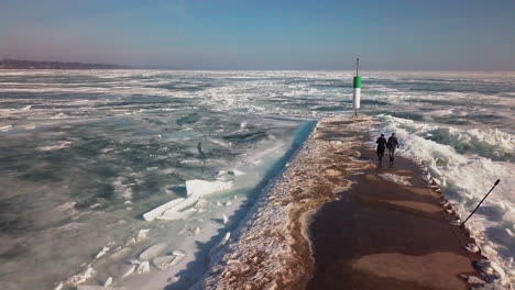 A-couple-holding-hands-walks-down-a-dock-on-a-frozen-lake-towards-a-lighthouse