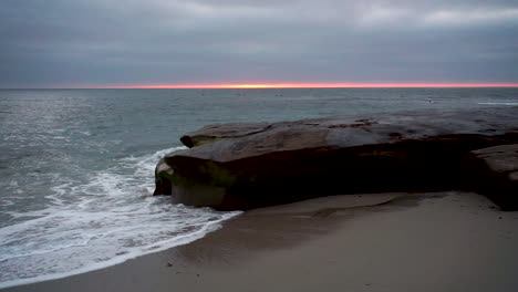 Die-Wellen-Krachen-An-Der-Küste-Von-San-Diego-Gegen-Einen-Felsen,-Wenn-Die-Sonne-Hinter-Dunklen-Wolken-Untergeht