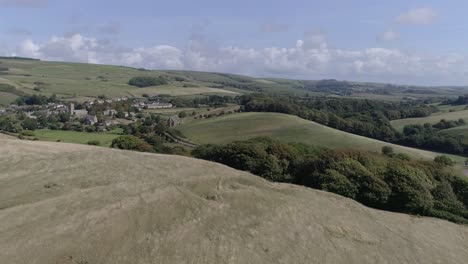 Low-tracking-aerial-moves-forward-fast-up-above-a-hill-to-reveal-the-village-of-Abbotsbury,-in-the-Dorset-countryside
