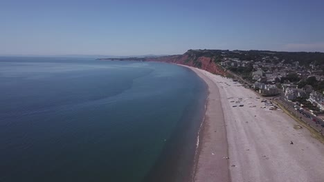 Wide-aerial-view-of-a-blue-sea,-pebble-beach,-rusty-red-Triassic-cliffs-and-tranquil-town-of-Budleigh-Salterton