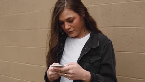 Steadicam-shot-of-a-beautiful,young,-brunette-college-teenager-texting-on-a-cell-phone-wearing-a-white-shirt-with-a-black-jacket-against-a-wall