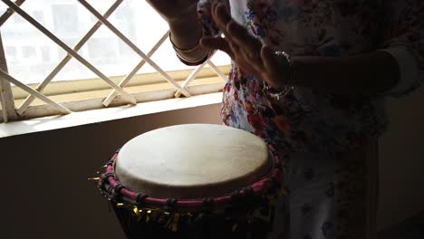 Medium-wide-looking-down-isolated-shot-of-a-woman-playing-djembe-drum-by-the-side-of-a-window
