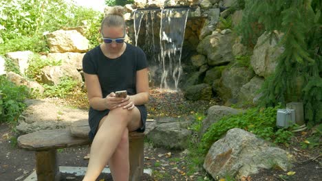 Tall-Woman-In-Short-Black-Dress-Sits-On-A-Bench-Near-A-Waterfall,-Tilting-Reveal-Of-An-Attractive-Girl-In-A-Park-Using-Her-Smart-Phone-To-Text-Her-Friends