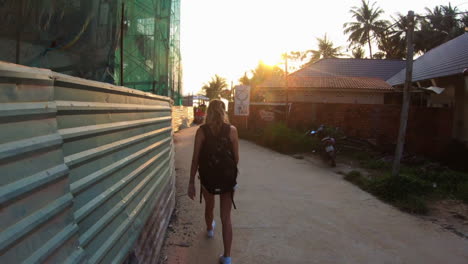 blonde-girl-wearing-backpack-walking-towards-the-sunset-and-flicking-her-hair-back-towards-the-camera