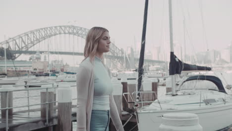 Junge-Schöne-Frau,-Die-Auf-Einem-Kai-Mit-Yachthafen-Und-Sydney-Harbour-Bridge-Hintergrund-In-Zeitlupe-Positiv-Und-Friedlich-Spaziert