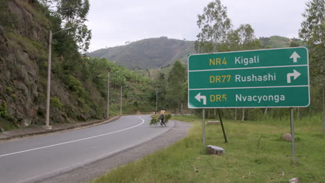 Pan-right-to-road-sign-leading-to-Kigali,-Rwanda-with-person-on-a-bike-riding-through-the-frame