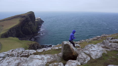 Tracking-follow-shot-of-a-girl-walking-on-the-edge-with-cliffs-in-foreground-and-Neist-Point-Lighthouse-and-Atlantic-Ocean-in-the-background-trying-to-take-a-photograph-in-Scotland,-Isle-of-Skye