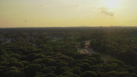 a-slow-aerial-dolly-shot,-pull-away-over-the-green-tree-tops,-in-a-park-at-sunset,-as-a-plane-flies-left-in-the-distance---car-traffic-on-the-highway-to-the-right