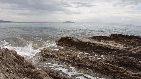 A-landscape-shot-of-waves-splashing-on-a-rocky-coast-in-Greece-on-a-sunny-day