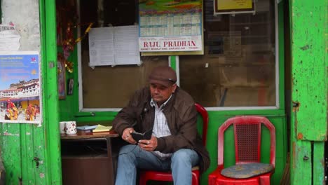 Un-Hombre-Indio-De-40-Años-Sentado-En-Una-De-Las-Tiendas-En-El-Mercado-Callejero-De-Darjeeling-Y-Revisando-Su-Teléfono