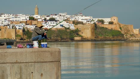 Anciano-Pescando-En-Primer-Plano-Con-La-Antigua-Fortaleza-En-El-Fondo