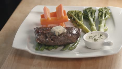 Mittlere-Schiebeaufnahme-Eines-Saftigen-Steaks-Mit-Butter-Darauf-Und-Seiten-Mit-Karotten,-Brokkoli-Und-Grünen-Bohnen-Auf-Einem-Hölzernen-Küchentisch