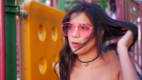 A-young-hispanic-woman-hipster-wearing-vintage-fashion-clothing-and-retro-pink-aviator-sunglasses-in-a-park-playground-SLOW-MOTION