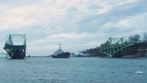 Small-fishing-ship-enters-the-port-via-an-closing-Oskara-Kalpaka-swing-bridge-in-Liepaja-in-cloudy-autumn-day,-wide-shot