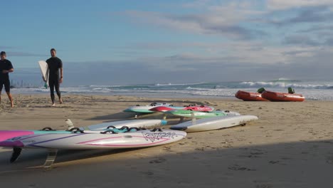 Surfbretter-Und-Rettungsboote-Liegen-Auf-Dem-Strandsand---Zwei-Aktive-Surfer,-Die-Nach-Dem-Surfen-Am-Strand-Spazieren-Gehen---Currumbin,-Gold-Coast,-Australien