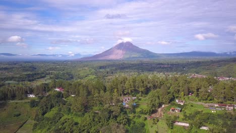 Reverse-drone-shot-of-Mount-Sinabung-volcano-and-the-surrounding-beautiful-green-village-in-Berastagi,-North-Sumatera-Indonesia