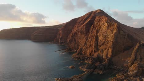 Sensational-dramatic-flight-approaching-beautiful-golden-rocky-rough-brown-mountain-peak-by-sandy-beach-of-Calheta-on-shoreline-coast-at-sunset,-Madeira,-Portugal,-overhead-aerial-toward