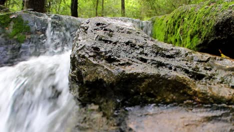 Wide-angle-close-up-view-of-a-large-wet-rock-with-a-waterfall-behind-it-to-the-left-and-another-fully-moss-covered-rock-on-the-right
