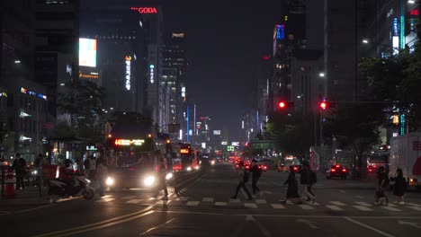 Busy-urban-city-street-life-with-pedestrians-crossing-the-road-by-buses-and-traffic-at-night,-static,-Seoul,-South-Korea