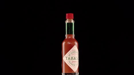 Static-Slow-Motion-Shot-Of-A-Tabasco-Bottle-Turning-On-Itself-With-Red-Chili-Peppers-Poping-Up-Behind-it,-Shoot-in-a-Studio-with-Black-Background