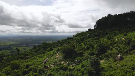 Phnom-Kulen,-Cambodia's-lush-green-countryside-low-fly-over-hill-with-sandstone-rock-formations-with-cloudy-skyline-during-rainy-season