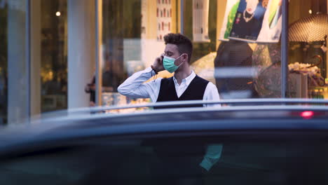 An-elegant-young-guy-wearing-a-simple-green-protective-Covid-19-facemask,-a-black-waistcoat,-white-shirt-and-jeans-standing-in-front-of-a-high-end-shop,-making-an-important-phone-call,-static-4k