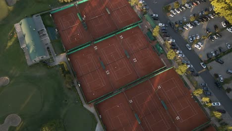 Aerial-high-angle-view-revealing-tennis-courts-between-a-golf-course-and-a-parking-lot-on-a-sport-club-at-sunset