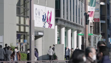 People-Wearing-Facemask-Walking-At-Street-On-A-Sunny-Day-With-Cancelled-Tokyo-2020-Olympic-Banner-Display-On-Exterior-Of-Building-During-Pandemic-In-Tokyo,-Japan