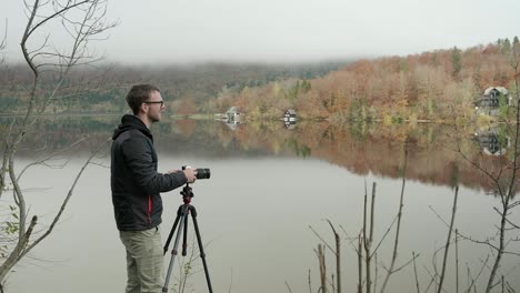 Mann-Organisiert-Seine-Kamera,-Um-Ein-Bild-Einer-Wunderschönen-Seespiegelung-Und-Perfekter-Herbstfarben-Im-Hintergrund-Und-Tief-Liegendem-Nebel-Im-Hintergrund-Aufzunehmen