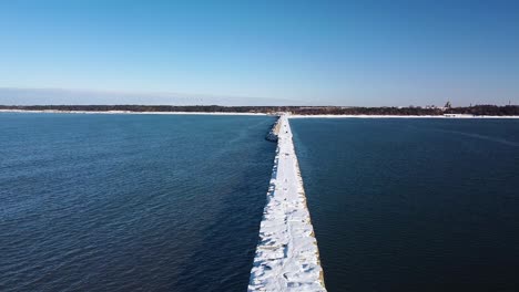 Aerial-view-of-snow-and-ice-covered-concrete-pier-in-the-calm-Baltic-sea,-Port-of-Liepaja-on-a-sunny-winter-day,-wide-angle-ascending-drone-shot
