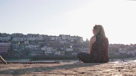 Back-View-Of-A-Blonde-Girl-Sitting-On-The-Quay-Looking-In-The-Distance-With-St-Ives-In-The-Background-On-A-Sunset-in-Cornwall,-England,-UK
