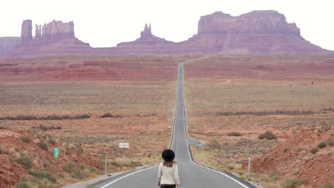 Junge-Frau-Mit-Hut-Läuft-Auf-Der-Berühmten-Leeren-Straße,-Die-Zu-Den-Sandsteinkuppen-Des-Monument-Valley-Navajo-Tribal-Park-An-Der-Grenze-Zwischen-Arizona-Und-Utah-In-Den-USA-Führt-–-Zeitlupenkamera-Neigt-Sich-Nach-Oben