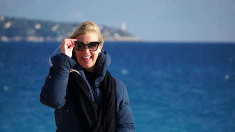 A-young-blonde-woman-laughs-to-the-camera-and-puts-her-sunglasses-on,-the-blue-sea-in-soft-focus-behind-her