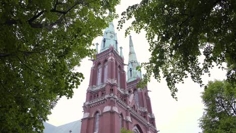 Static-shot-of-St-Johns-Church,-filmed-under-trees,-green-leaves-and-branches,-waving-in-the-wind,-on-a-cloudy,-summer-day,-in-Ullanlinna,-Helsinki,-Finland