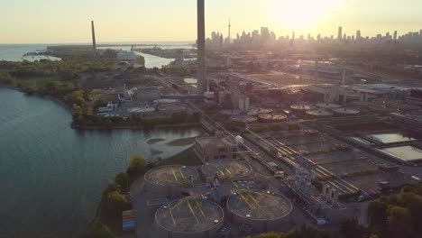 Aerial-Sunset-Wide-Shot-Sliding-Left-Over-Lake-Shoreline-Showing-Industrial-Factory-And-Huge-Smokestack-Tower-With-Downtown-Skyline-In-Background-In-Toronto-Ontario-Canada
