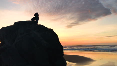 Silhouette-of-two-girls-on-a-rock-at-Bandon-beach,-one-taking-photos-of-the-sunset