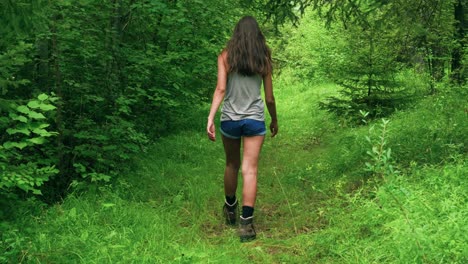A-tall-brunette-woman-walking-down-a-green,-grassy-forest-path-lined-with-tall-trees-into-the-distance