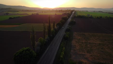 Luftaufnahme-Von-Autos,-Die-Bei-Sonnenuntergang-Im-Sommer-Auf-Einer-Straße-In-Der-Nähe-Von-Feldern-Entlangfahren