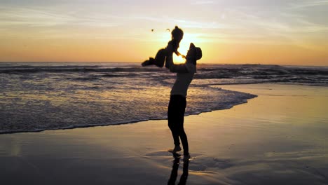 Eine-Mutter-Genießt-Den-Urlaub-Mit-Ihrem-Kind-Bei-Sonnenuntergang-Am-Strand,-Während-Sie-Ihr-Kind-Vor-Freude-In-Die-Luft-Wirft