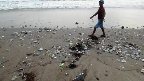 Einheimische-Gehen-Auf-Bali-Am-Ufer-Entlang,-Während-Wellen-Plastikmüll-Aus-Dem-Meer-Spülen,-Verschmutzte-Küste