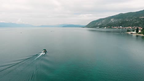 Toma-Aérea-Siguiendo-El-Barco-En-El-Lago-Ohrid-En-Mcedonia-Con-Vista-A-La-Costa,-Al-Acantilado-Y-Al-Hermoso-Agua-Azul