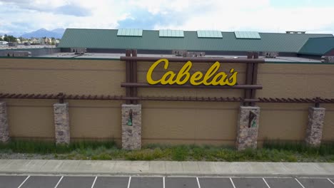 Aerial-footage-of-the-sign-in-the-Cabela's-parking-lot-in-Anchorage,-Alaska-also-showing-many-recreational-vehicles-parked-with-birds-flying-and-the-sun-shining-on-the-mountains-in-the-background